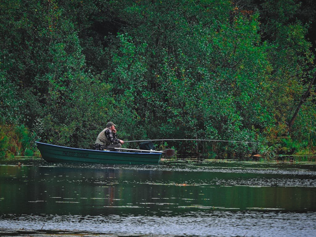 A man enjoys fishing from a boat on a serene lake surrounded by lush greenery.